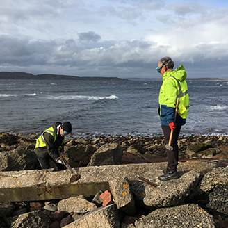 A researcher in high-vis clothing uses a geology tool to explore discarded concrete on a beach as another researcher looks on