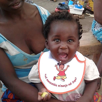 African mother holding her smiling baby on her lap. The baby is wearing a bib with a message on it promoting hygiene.