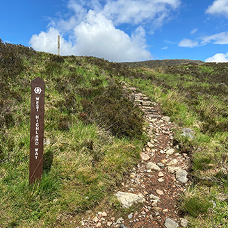 A rocky pathway leading up a grassy hill, with a sign saying the West Highland Way on the left  