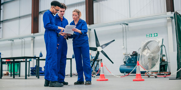 A team of researchers in a hangar wear blue jumpsuits and look at paperwork together