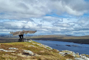 A large Golden Eagle statue overlooks a Hydro Electric Dam, and  turbines of a windfarm can be seen in the background.