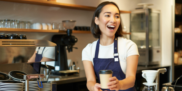 A barista smiling and serving a takeaway hot drink.