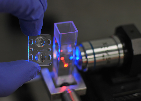 A hand holding a small 3D manufactured micro optic next to a laser shining through a small glass rectangular tube