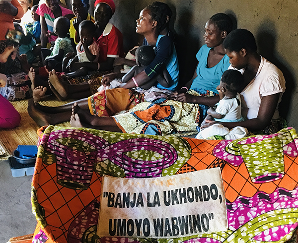 A colourful African bag in the foreground with mothers and children sitting against a wall behind it