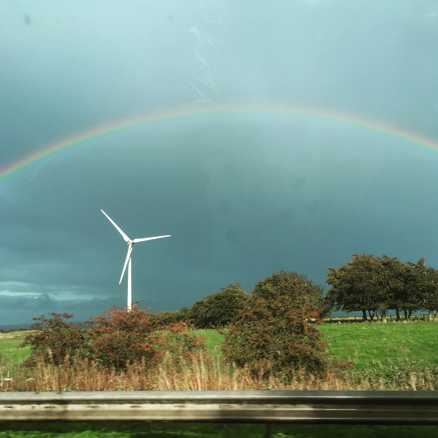 A single wind turbine on a hillside with a rainbow arching over it