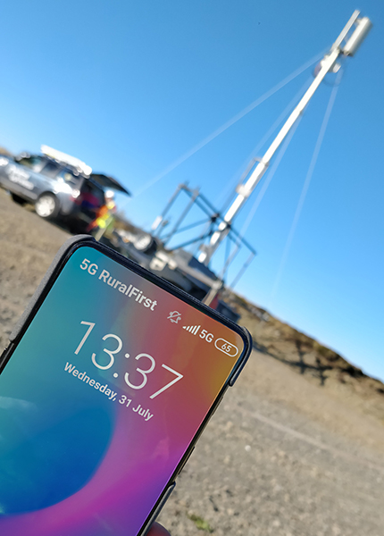 A hand holding a mobile phone in front of a phone mast in a remote location