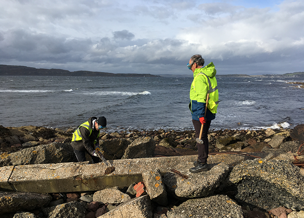 A researcher in high-vis clothing uses a geology tool to explore discarded concrete on a beach as another researcher looks on