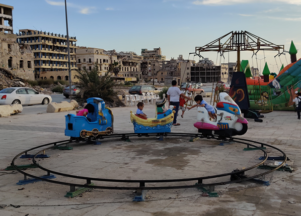 Young children play in a makeshift fun fair with bombed buildings in the background 