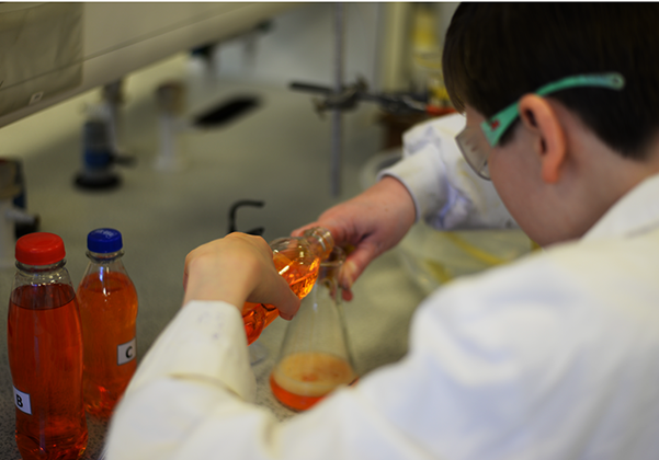 A child in a lab coat pouring an orange liquid into a beaker
