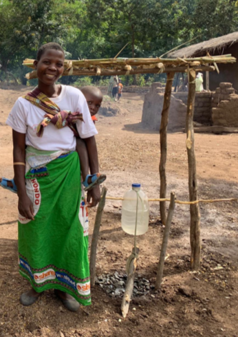 An African woman with a young child on her back standing next to an outdoor kitchen area