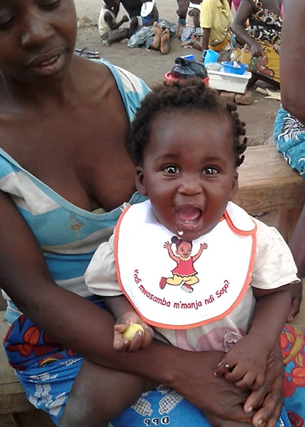 African mother holding her smiling baby on her lap. The baby is wearing a bib with a message on it promoting hygiene