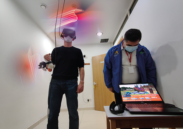 A man stands wearing a VR headset and holding a controller, next to a man looking at a game screen on a laptop