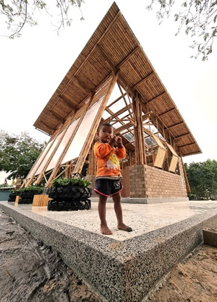 A child stands in front of a bamboo building