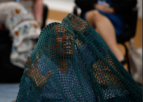 An African woman kneels with hands in the air as if in pain, covered with a large blue fishing net, surrounded by an audience