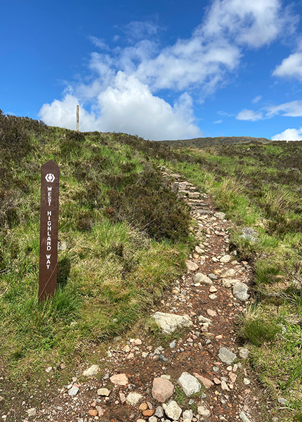 A rocky pathway leading up a grassy hill, with a sign saying the West Highland Way on the left  