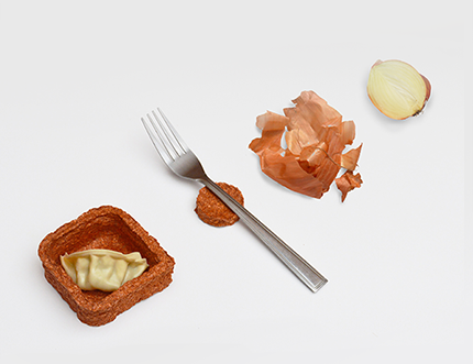 A slice of onion next to crumpled onion skins, a fork next to a little rough-textured container holding a gyoza