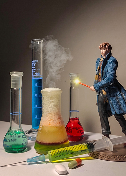 A figurine of a man with a wand stands to the right of various beakers filled with colourful liquids with smoked emerging, and a syringe and tablets in the foreground.