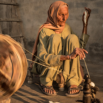 An elderly lady sits in the street weaving on a traditional loom, in rural India