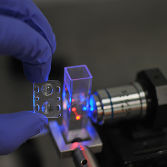 A hand holding a small 3D manufactured micro optic next to a laser shining through a small glass rectangular tube