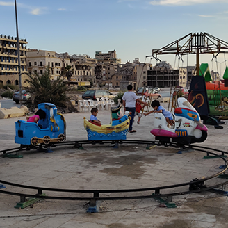 Young children play in a makeshift fun fair with bombed buildings in the background 