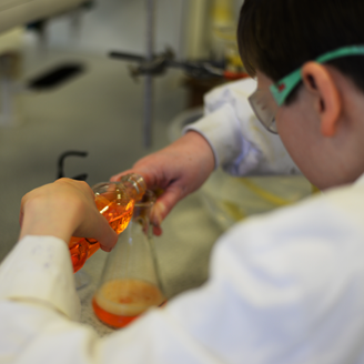 A child in a lab coat pouring an orange liquid into a beaker