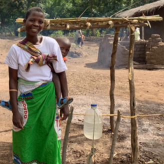 An African woman with a young child on her back standing next to an outdoor kitchen area