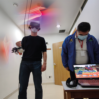 A man stands wearing a VR headset and holding a controller, next to a man looking at a game screen on a laptop