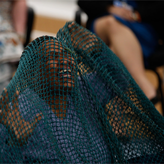 An African woman kneels with hands in the air as if in pain, covered with a large blue fishing net, surrounded by an audience