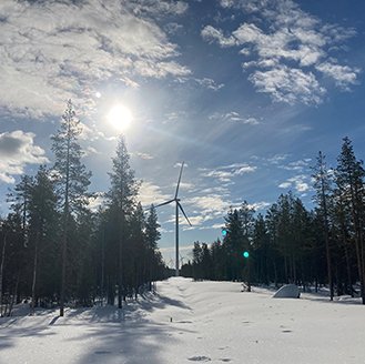 A snowy landscape with tall pine trees lining the horizon and a wind turbine in the middle. The sun low in the sky just above.
