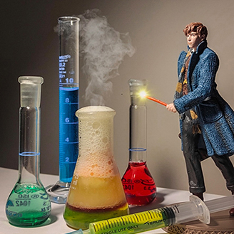 A figurine of a man with a wand stands to the right of various beakers filled with colourful liquids with smoked emerging, and a syringe and tablets in the foreground.