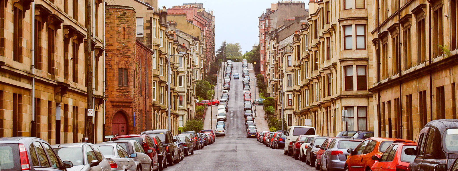 a view of a long Glasgow street filled with tenement flats with cars lined on both sides of the road