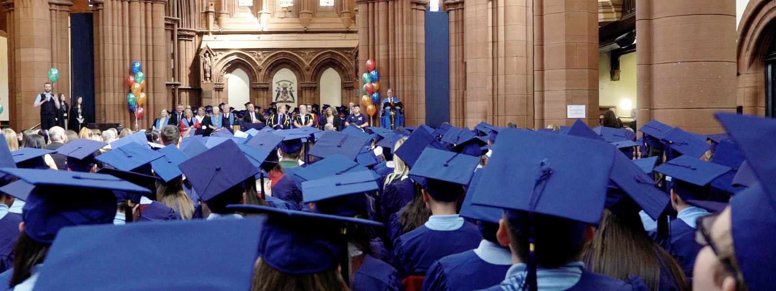 Pupils dressed in blue gowns and mortarboards sit in the Barony Hall facing the stage.