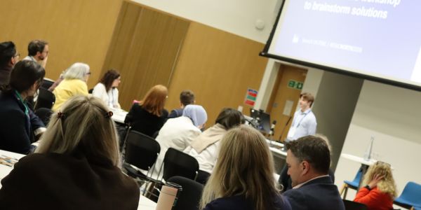 A group of conference delegates sitting at tables in an auditorium, while a speaker stands under a presentation screen.