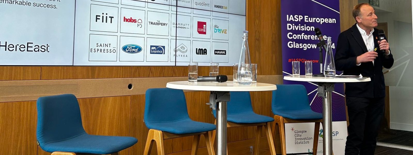 A keynote speaker stands in front of a videowall, holding a mic, delivering a conference speech