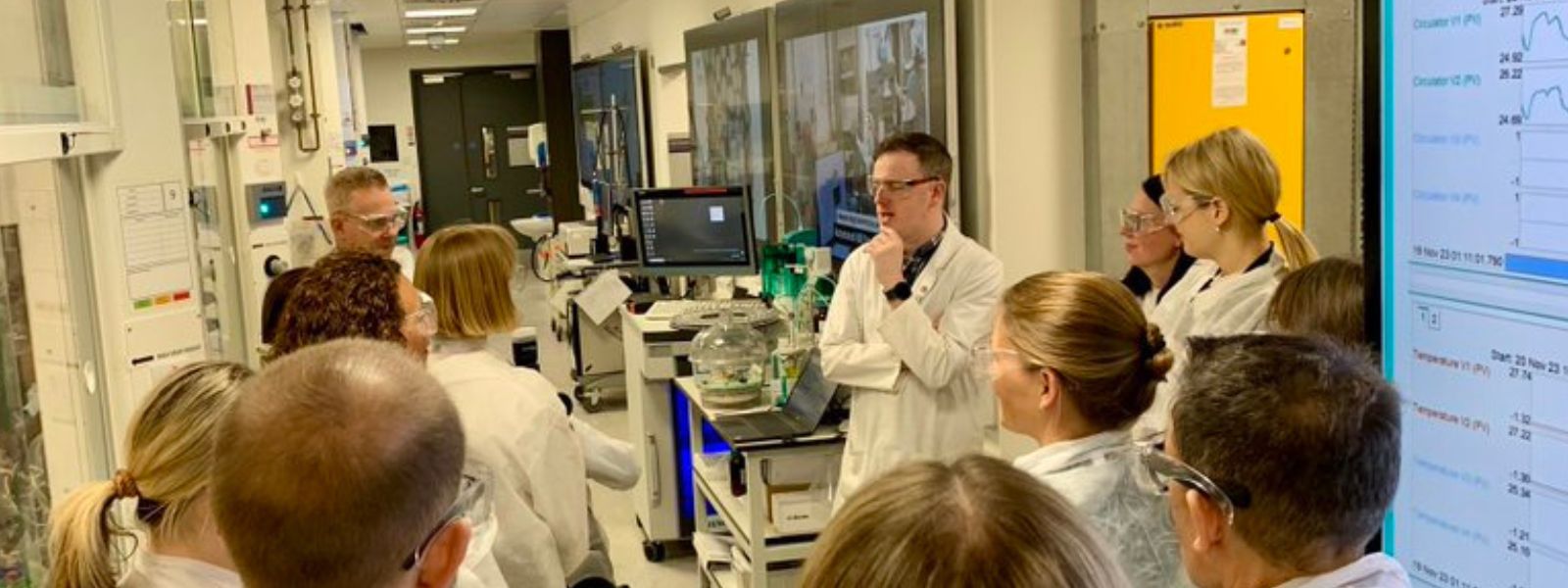 A group of people wearing white coats stand in a science laboratory