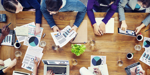 a birdseye view of a team sitting and working around a table with charts, ipads and papers strewn across the table