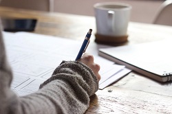 A hand writing on paper with a mug in the background