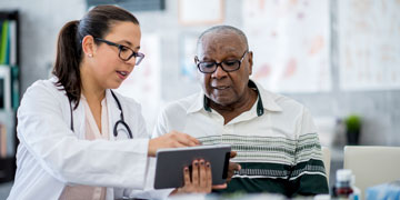 A senior male patient is indoors in a hospital room. He is watching his female doctor using a tablet computer. 