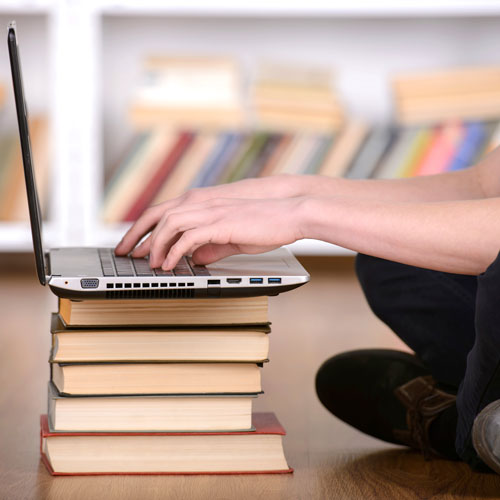 side profile of a person sitting cross legged on the floor, typing on a laptop which is being propped up by a pile of books