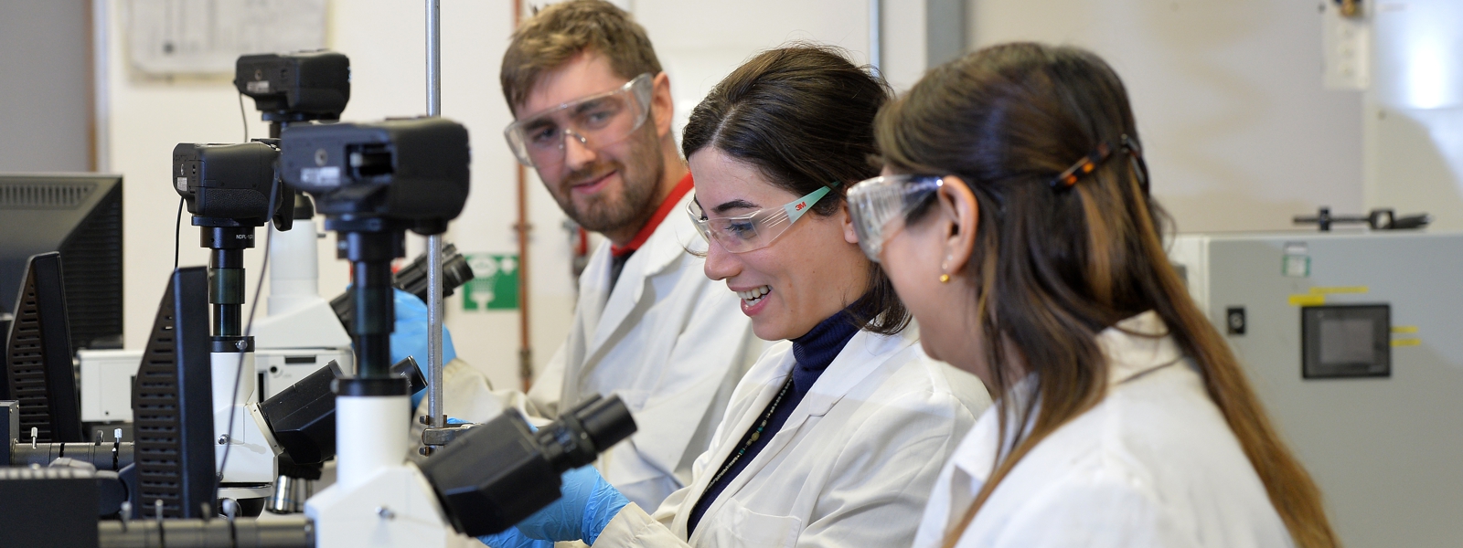 three postgraduate research students smile and work in a lab wearing lab coats