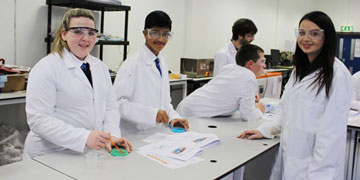 school pupils wearing lab coats and safety glasses smile at the camera during a lab experiment