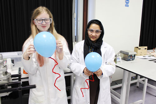 Two female school pupils wearing lab coats and safety glasses stand together holding balloons while smiling at the camera