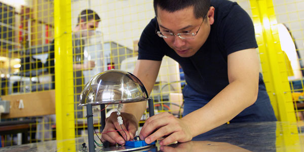 A scientist working in a high voltage lab, inside a yellow cage, attaching wires to a piece of equipment