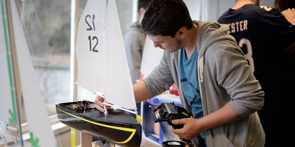 a student holds a remote control in his left hand, while tending to his hand made boat with his right hand