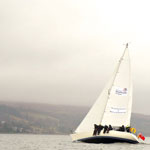 catalina yacht in the water on a grey and mistry day, tipping to the right, while it's passengers sit with legs dangling over the edge