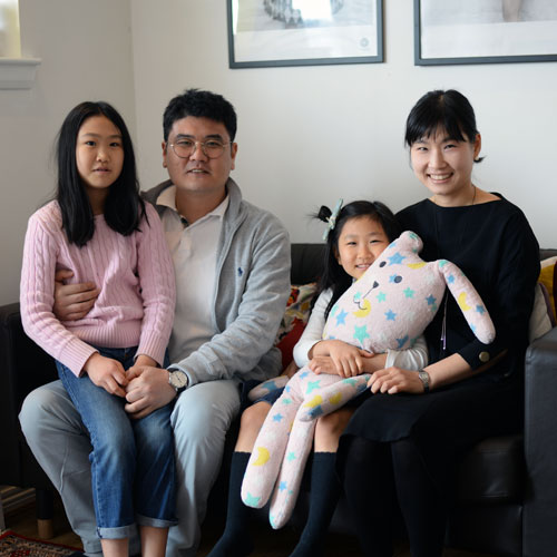Naval Architecture student Sung il sits with his wife and their two daughters in their living room and smile at the camera