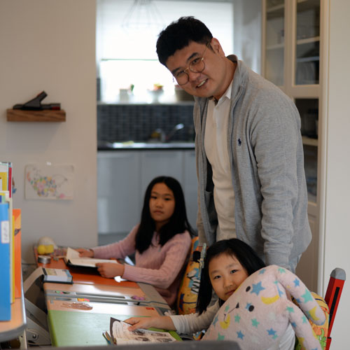 Sung il with his daughters as they sit at their desks in their living room, with Sung il standing behind them, all smiling at the camera
