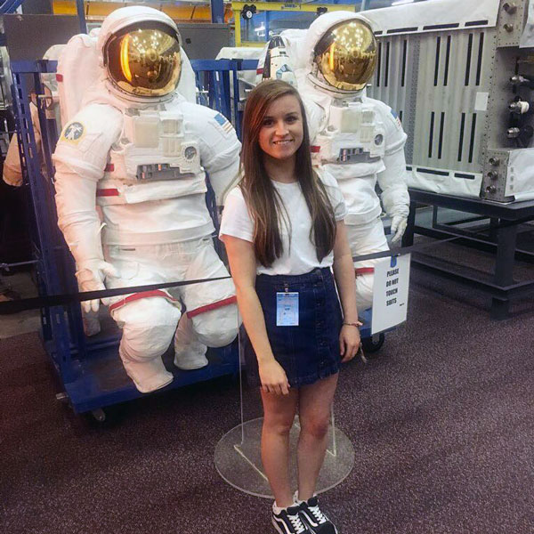 Engineering student Gemma standing in front of two astronaut suits, smiling at the camera