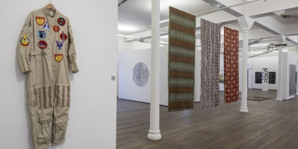 Exhibition at a gallery with a pale badged bodysuit costume on the left and three patterned coloured sheets hanging on the right