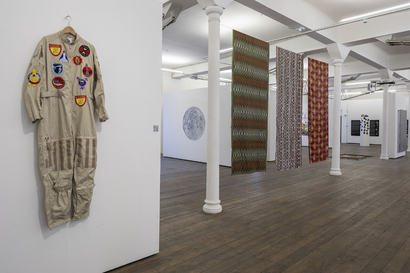 Exhibition at a gallery with a pale badged bodysuit costume on the left and three patterned coloured sheets hanging on the right